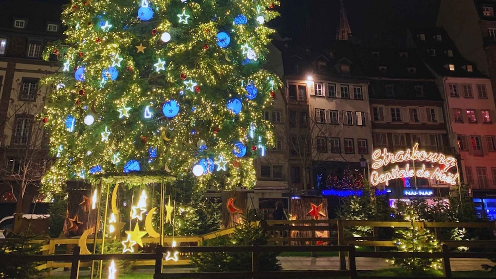 A large decorated Christmas tree with lights and ornaments in a public square, surrounded by buildings and a neon sign. 1