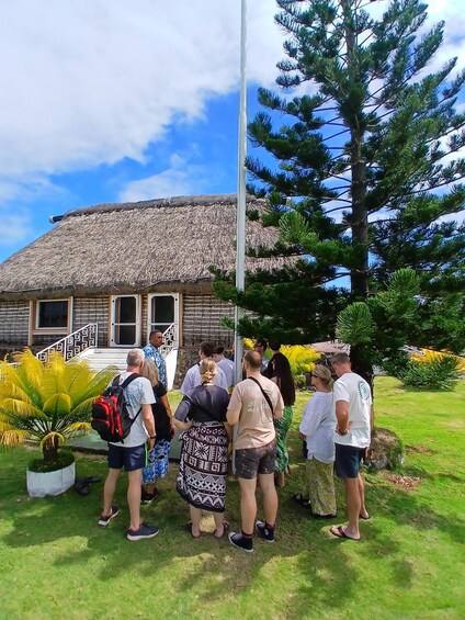 A group of people standing in front of a thatched-roof building with a flagpole nearby. 3