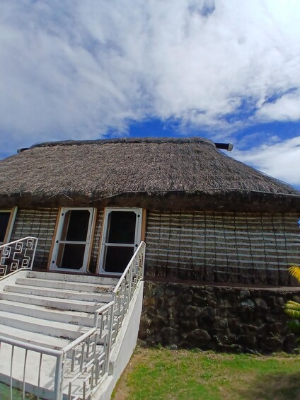 A traditional thatched-roof house with a stone wall, a wooden staircase, and a metal railing. 1