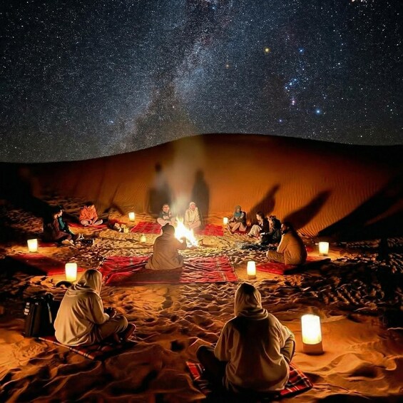 A group of people sitting on a sand dune at night under a starry sky, with a fire in the center. 1