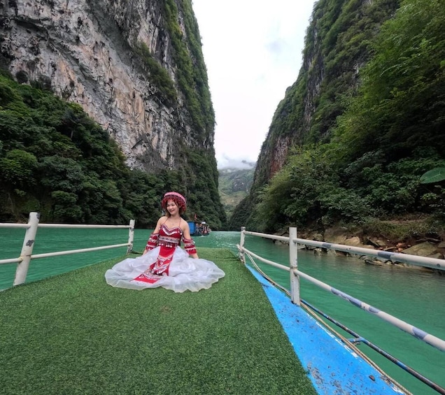 A person in traditional attire sitting on a bridge with a river and steep cliffs in the background. 1