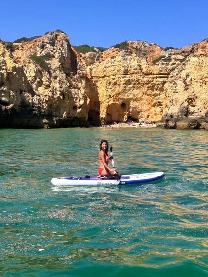 A person sitting on a paddleboard in clear water with rocky cliffs in the background. 2