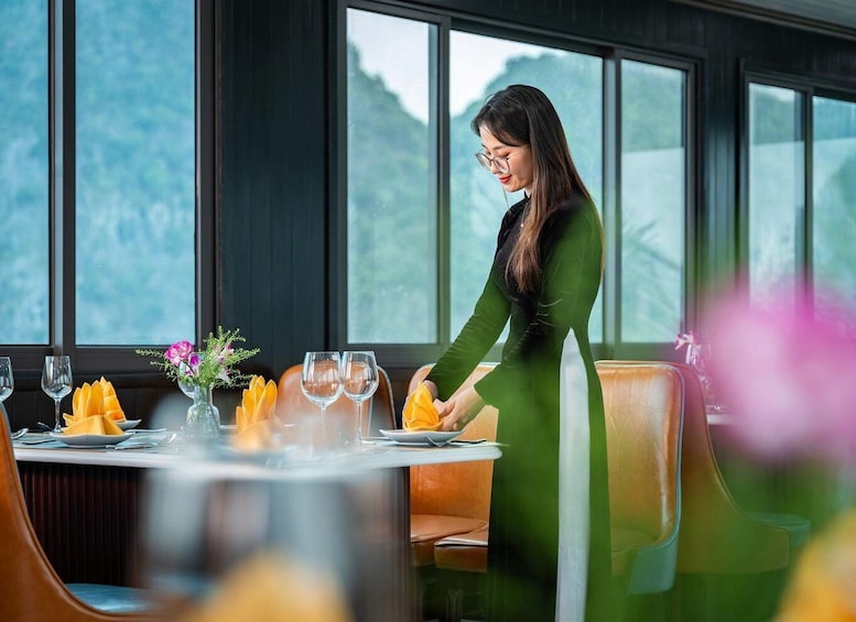 A woman in a green dress serving food at a restaurant with large windows and orange chairs. 5
