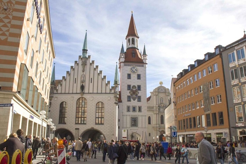 A busy urban square with historic buildings, a prominent clock tower, and a crowd of people. 5