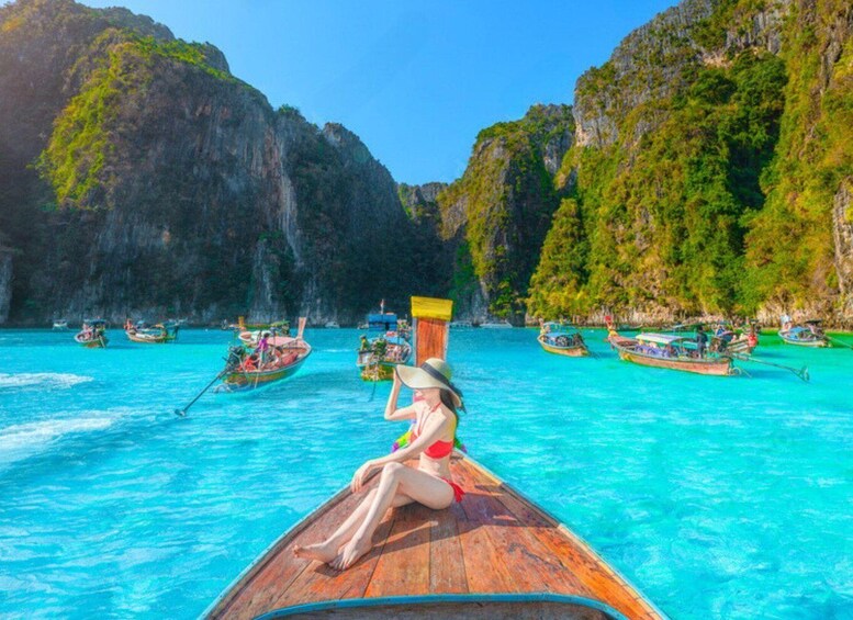 Een vrouw in een bikini zit op een houten boot in turquoise water, omringd door traditionele boten en weelderige kliffen. 1