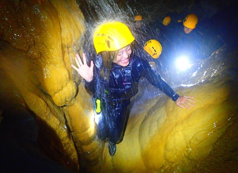 A person in a yellow helmet and black wetsuit inside a cave with yellow walls and water flowing down. 1