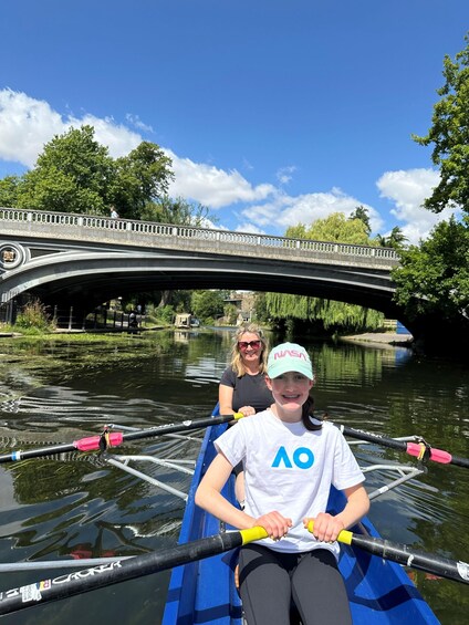 Two people rowing a blue boat on a river with a bridge in the background. 1