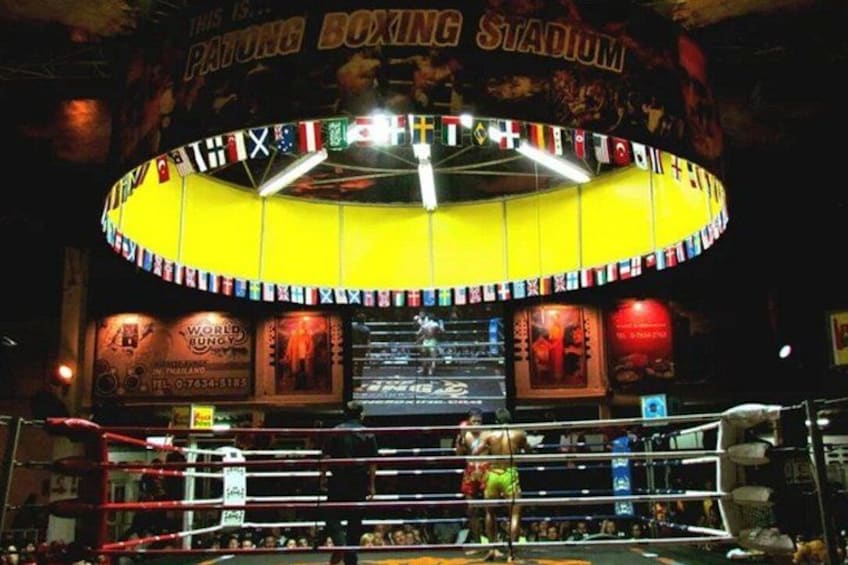 A boxing ring with a circular ceiling adorned with flags, a boxing poster, and a sign reading 'PATONG BOXING STADIUM.' 3