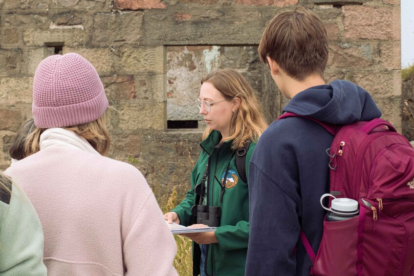 Three people standing outdoors, one holding a map, in front of a stone wall with a small window. 3
