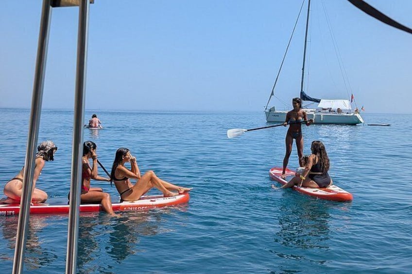 People on paddleboards in the ocean, a sailboat in the background, and calm water. 2