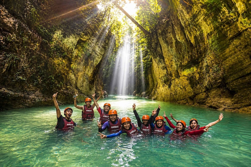 A group of people in a clear, turquoise river with a waterfall in a rocky, forested gorge. 3