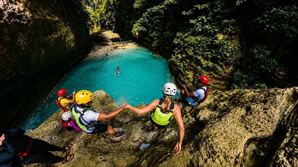 A group of people rappelling down a rocky cliff into a turquoise pool surrounded by lush greenery. 1