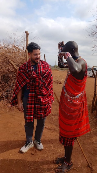 Two people in a rural setting, one wearing a red and black checkered garment and the other in a red patterned outfit, with a vehicle and sparse vegetation in the background. 5