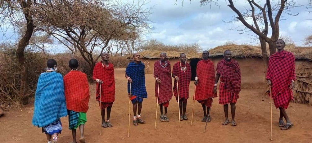 A group of people in traditional attire standing in a rural setting with trees and a thatched-roof structure. 1