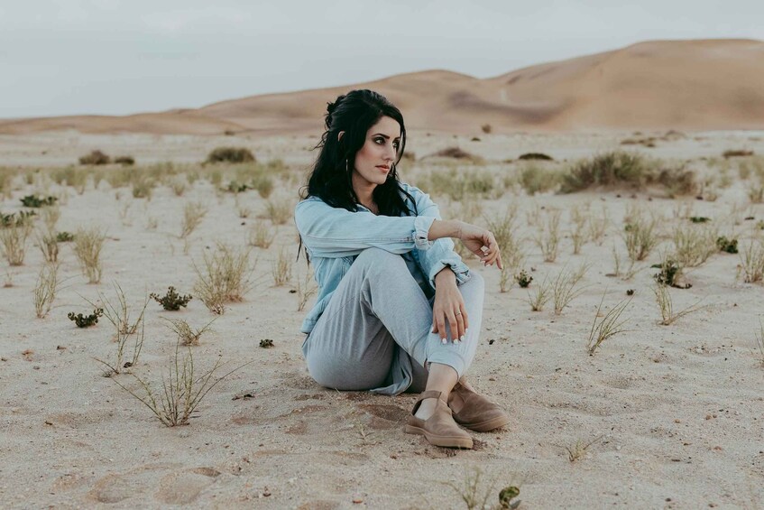 A person sitting on the sand in a desert landscape with sparse vegetation and sand dunes in the background. 5