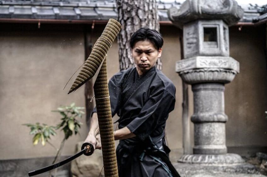 Eine Person in traditioneller Kleidung hält ein langes, gebogenes Objekt, möglicherweise eine Waffe, in einem Innenhof mit einer Stein Säule und einem Baum. 2
