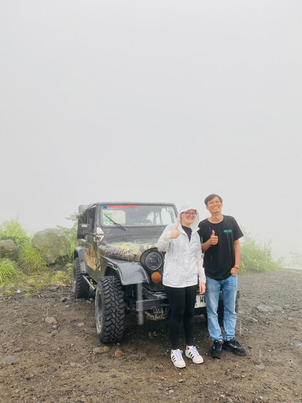 Two people standing next to a large off-road vehicle on a dirt path with vegetation in the background. 3