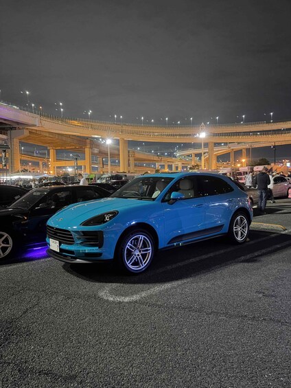 A blue car parked in a lot with a multi-level overpass in the background. 2