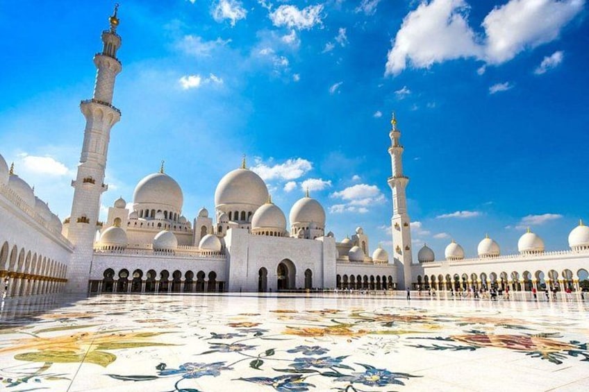 Un'ampia moschea con numerose cupole e minareti, un cortile spazioso caratterizzato da elaborate decorazioni in piastrelle, e un cielo azzurro intenso. 1