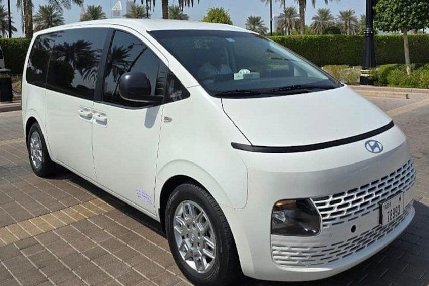 Un minivan bianco parcheggiato su una superficie pavimentata, con palme e un siepe sullo sfondo. 4