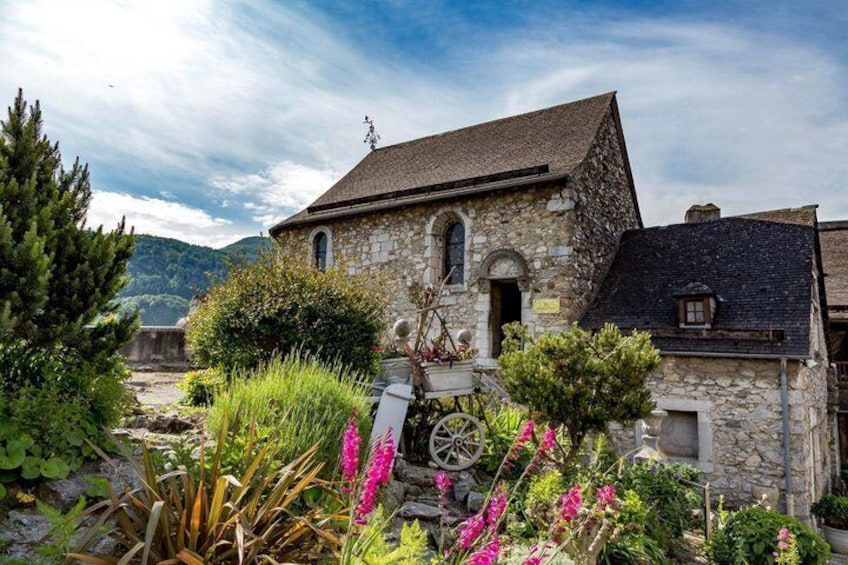 A stone building with a sloped roof, surrounded by lush greenery and flowering plants. 3