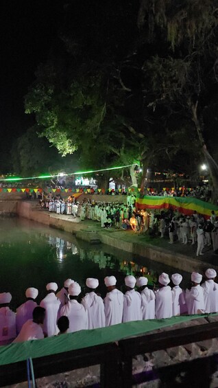 A nighttime outdoor gathering with people in white robes under large trees, a body of water, and green and red decorations. 1