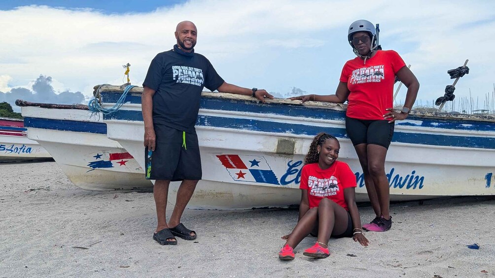 Three people standing and sitting near a boat with a beach in the background. 3