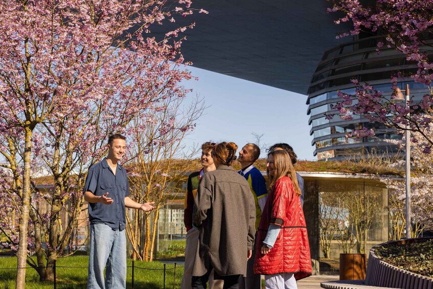 A group of people standing outdoors in a park with cherry blossom trees and a modern building in the background. 5