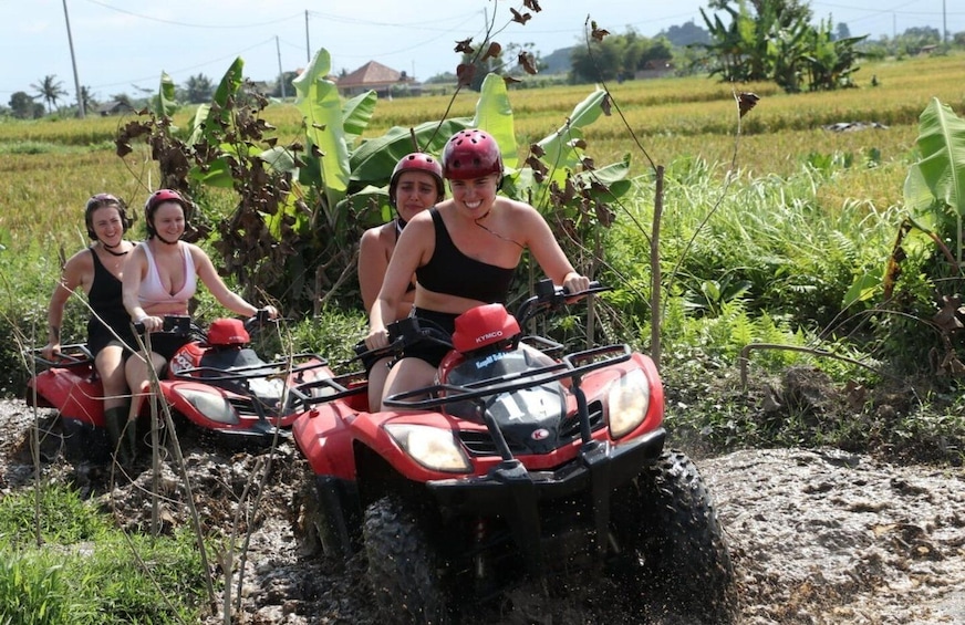 Four people on a red ATV in a muddy field with banana plants and a rural landscape in the background. 5