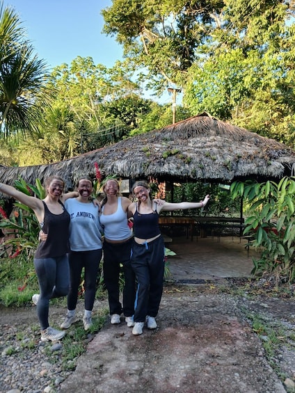 Four people standing in front of a thatched-roof structure with a garden and trees in the background. 2