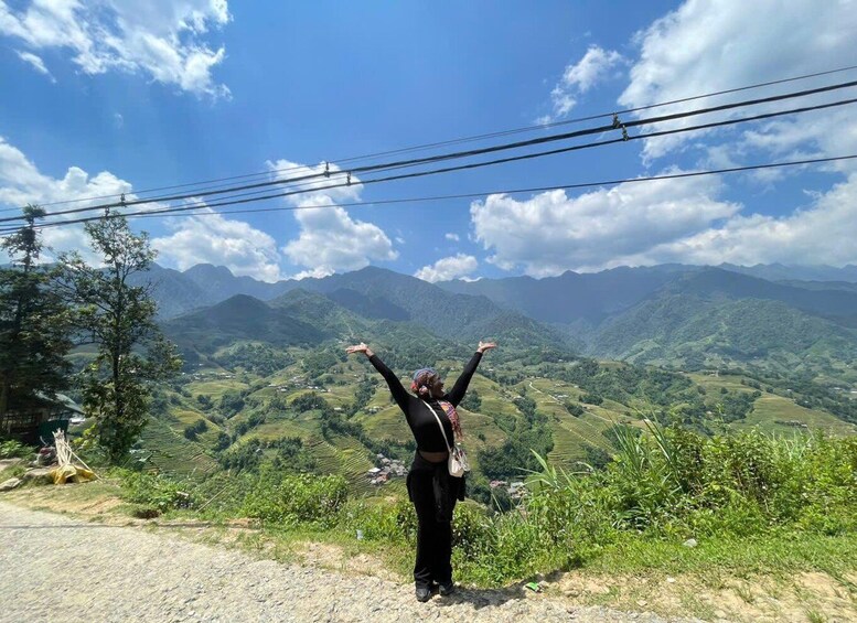 A person standing on a dirt path with arms raised, overlooking terraced fields and mountains under a clear sky. 5