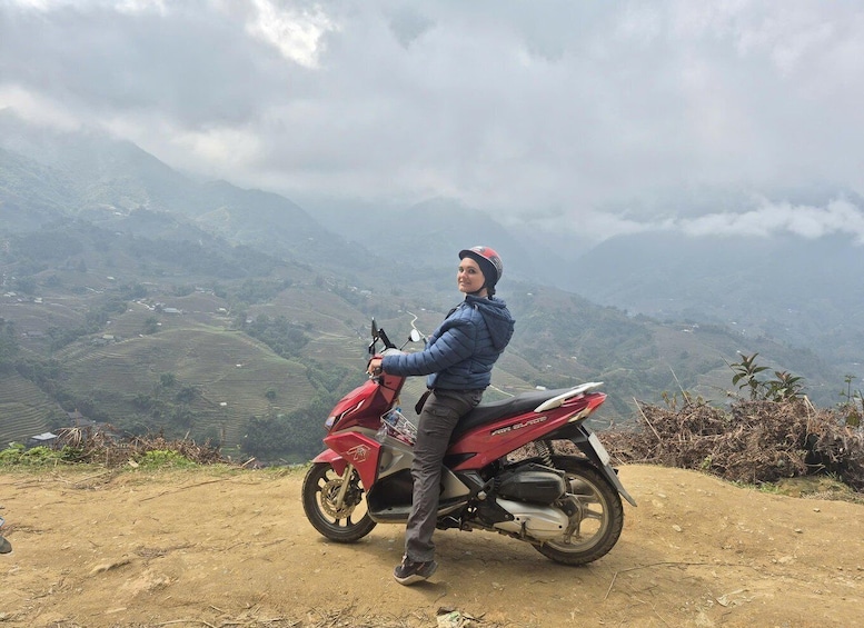A person sitting on a red motorcycle with a helmet, overlooking a hilly landscape with terraced fields. 2