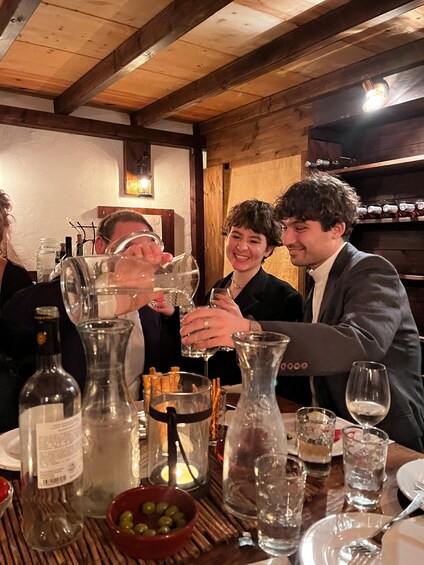 A group of people toasting with wine glasses at a wooden table with various bottles and glasses. 3