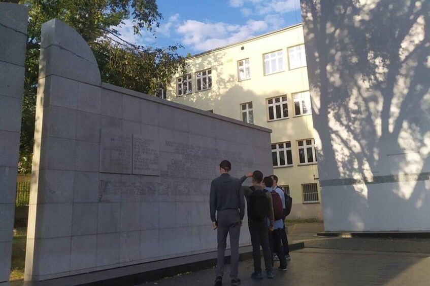 A group of people standing in front of a large stone wall with inscriptions, with a building in the background. 1