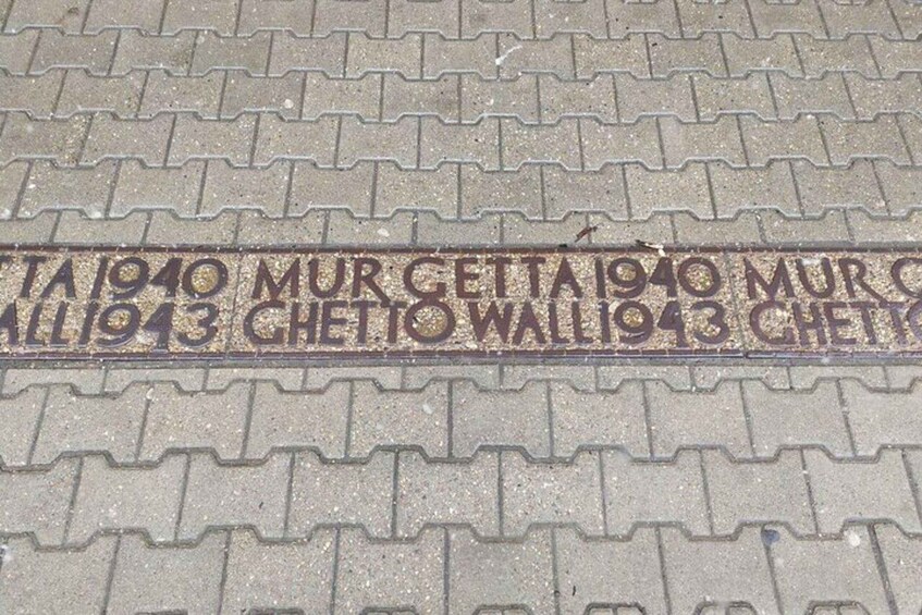 A brick pavement with engraved text reading "TA 1940 MUR GETTA 1940 MUR ALL 1943 CHETTO WALL 1943 CHETTO". 2