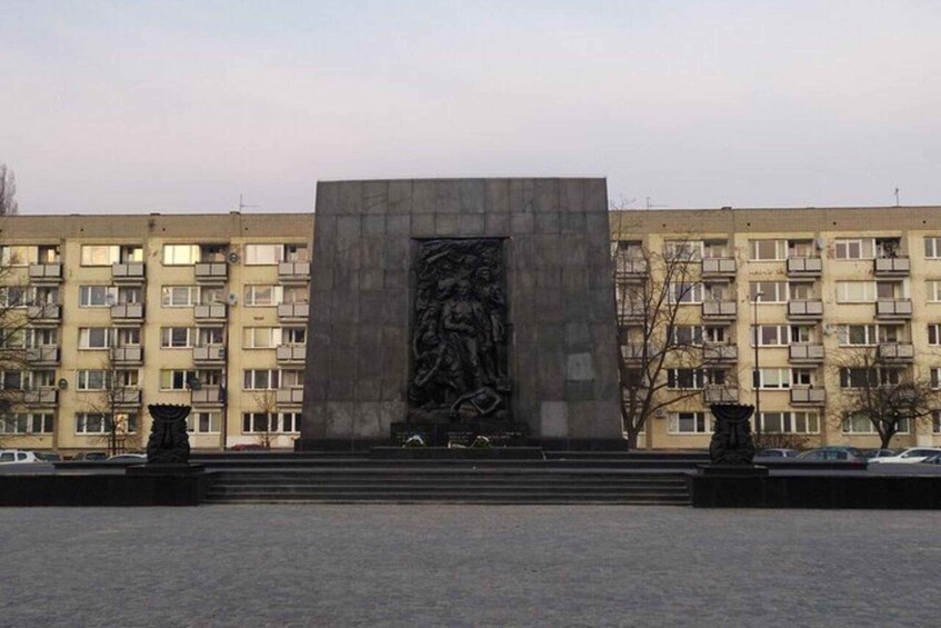A large, rectangular monument with a relief sculpture in the center, surrounded by a paved area and a multi-story residential building in the background. 5