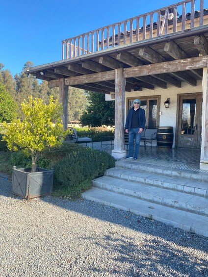 A man standing on a porch with a wooden railing, a potted tree, and a gravel pathway. 5