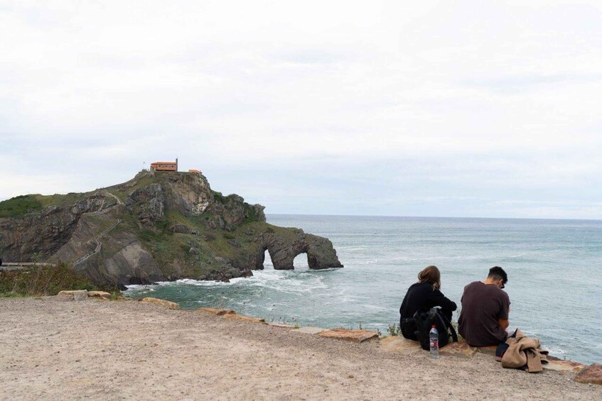 A rocky coastline with a small building on a hill and two people sitting by the sea. 1
