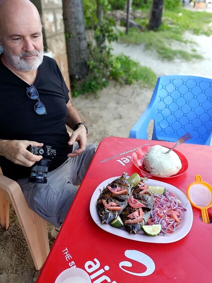 A man sitting at a table with a camera, a plate of food, and a blue chair. 2