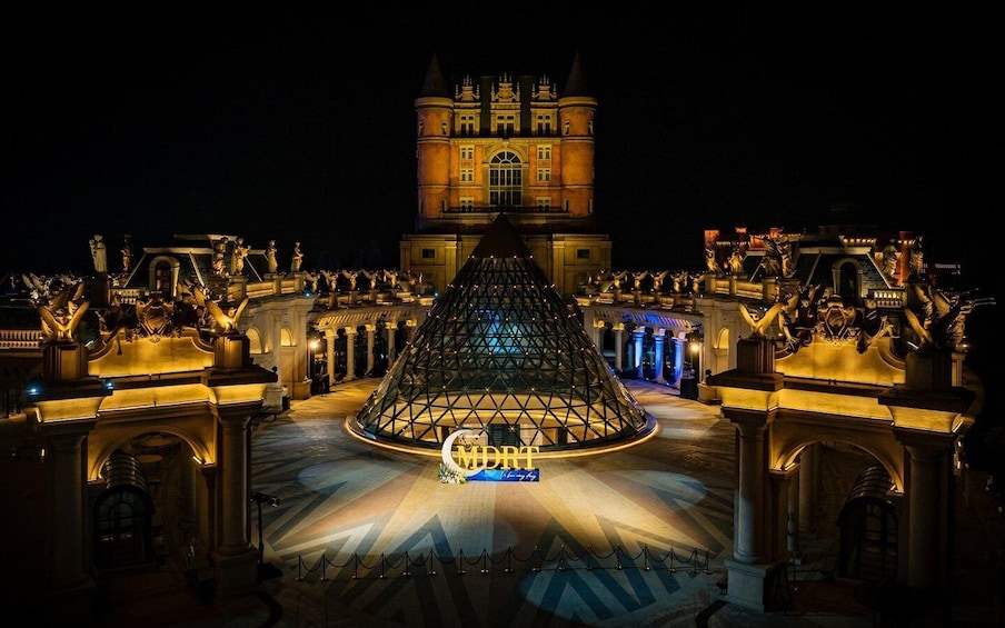 A grand illuminated building with a pyramid structure in the center, surrounded by statues and columns. 5