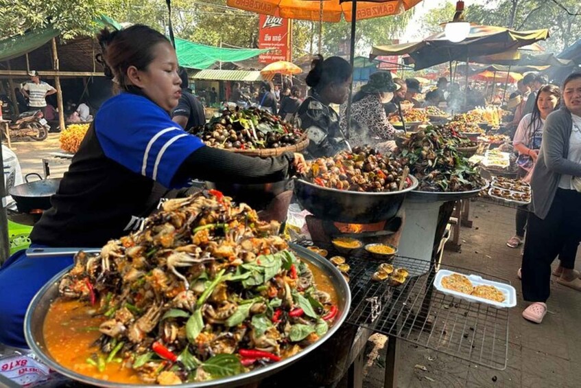 A street food market with large pots of grilled food and a woman serving food. 4