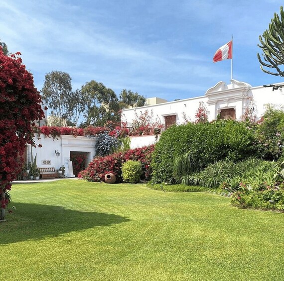 A white building with a red and white flag, surrounded by lush greenery and red flowering plants. 1