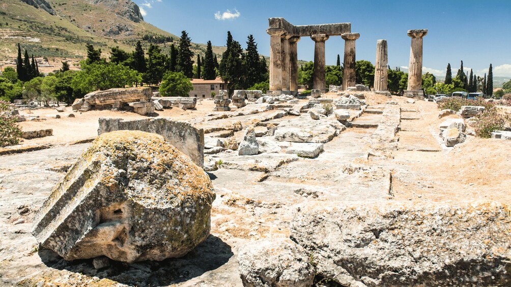 Ancient ruins with columns and stone blocks, surrounded by trees and a mountainous backdrop. 5