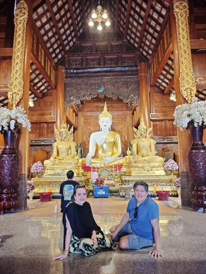 A large golden Buddha statue in a temple with intricate woodwork and two people sitting in front. 5
