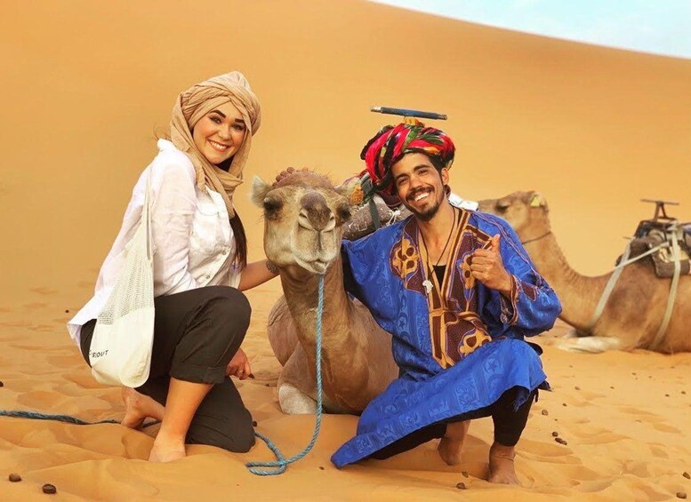 Two people in traditional attire with camels in a desert. 2