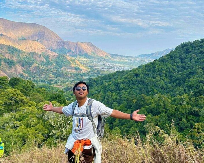 A person standing on a hill with arms outstretched, overlooking a valley with dense forest and mountains in the distance. 2