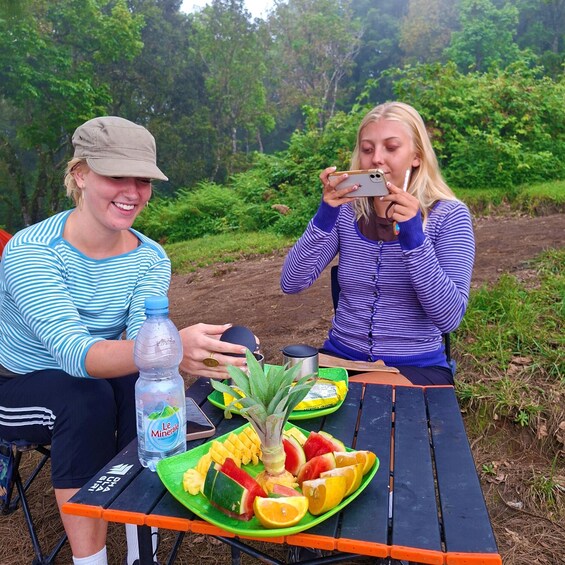 Two people sitting at an outdoor table with a plate of fruit and a water bottle. 3