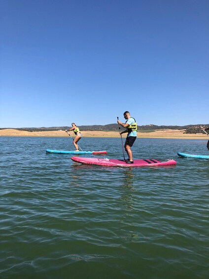 Two people paddleboarding on a lake with a clear sky and distant hills. 3