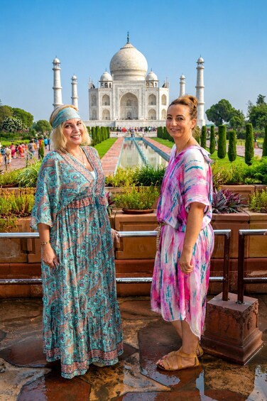 Two women standing in front of the Taj Mahal with a garden and a fountain in the background. 1