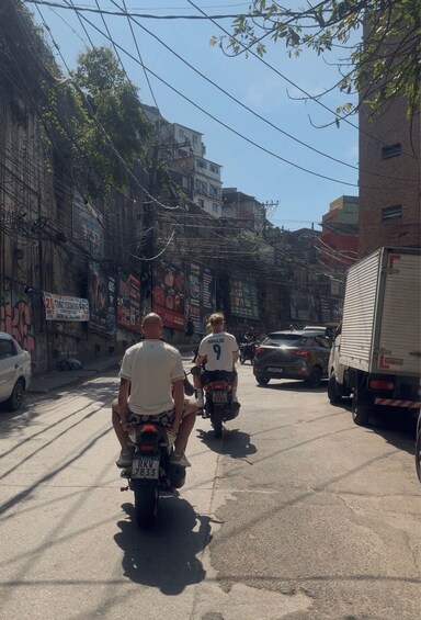 Two motorcyclists riding down a narrow urban street with buildings and power lines on either side. 4